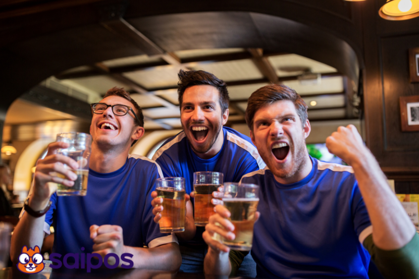 Três torcedores lado a lado, ambos de camiseta azul com listras brancas, segurando copos de cerveja. Todos sorriem e comemoram. À esquerda o logo da Saipos em azul. 