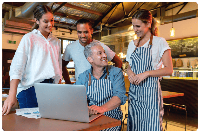 Quatro pessoas em um ambiente de restaurante moderno e iluminado. Um homem mais velho está sentado à mesa com um laptop aberto à sua frente, usando avental listrado azul e branco. Ele sorri enquanto interage com três colegas mais jovens — duas mulheres (uma com avental, outra com camisa branca) e um homem com camiseta branca — que estão em pé ao seu redor, também sorrindo. Ao fundo, há um balcão com vitrine de doces e cardápio na parede. A cena transmite colaboração e gestão de equipe em um restaurante.  Sistema para restaurantes. Saipos.