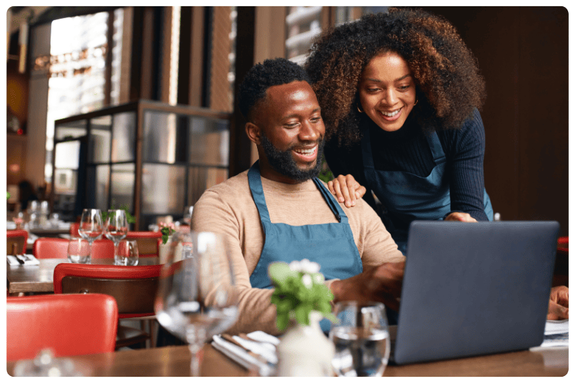 Duas pessoas com aventais olhando sorridentes para um notebook em um restaurante, representando o uso de tecnologia para controle de vendas para restaurantes. Saipos.