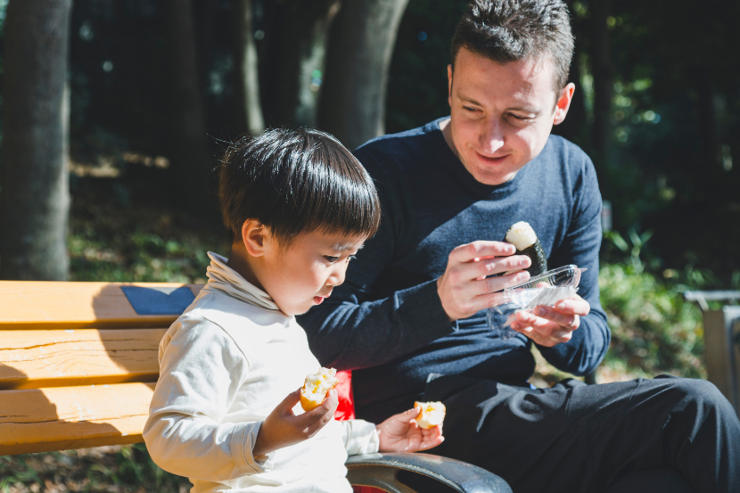menino e seu pai sentados ao ar livre em banco de madeira comendo comida japonesa