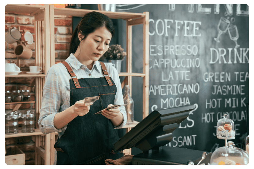 Mulher usando avental de barista conta dinheiro atrás do balcão de uma cafeteria. À sua frente há um computador de caixa e, ao fundo, um quadro-negro com o cardápio escrito com giz, mostrando bebidas como café, latte e cappuccino. O ambiente tem decoração acolhedora e organizada. SAIPOS
