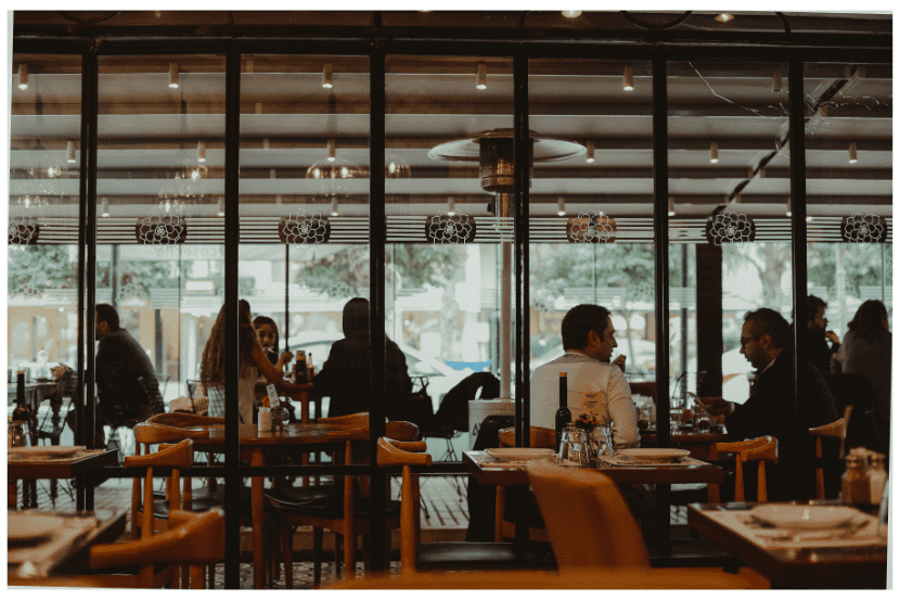 Restaurante visto através de uma parede de vidro com moldura preta. No interior, várias mesas de madeira estão ocupadas por clientes conversando e jantando. O ambiente tem luz amarelada e acolhedora, com luminárias pendentes no teto e aquecedores. Pessoas de diferentes idades interagem em clima descontraído. Foco no atendimento acolhedor e no ambiente organizado, ideal para quem busca um sistema eficiente de controle de vendas. SAIPOS