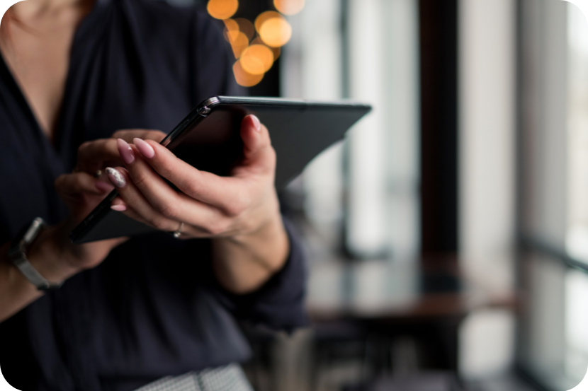Imagem mostra abaixo da cabeça de uma mulher com zoom nas mãos segurando o tablet cinza com unhas grandes e janelas desfocadas ao fundo