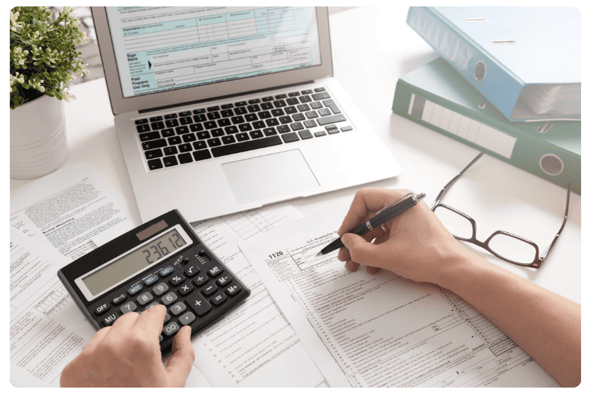 imagem de uma mesa com notebook, calculadora e papel. Há uma pessoa realizando uma conta.