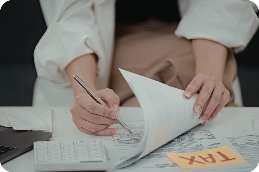 mesa branca com pilha de documentos sendo seguradas por mãos. Apenas os braços aparecem, com camiseta branca e uma caneta dourada sendo segurada.