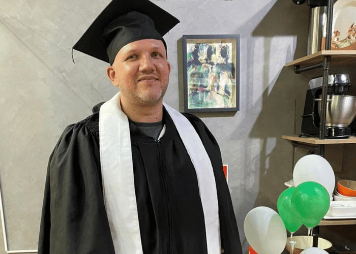 Homem sorridente vestindo beca e capelo de formatura preto, com quadro e estante ao fundo e balões coloridos decorativos à direita, celebrando sua conquista acadêmica