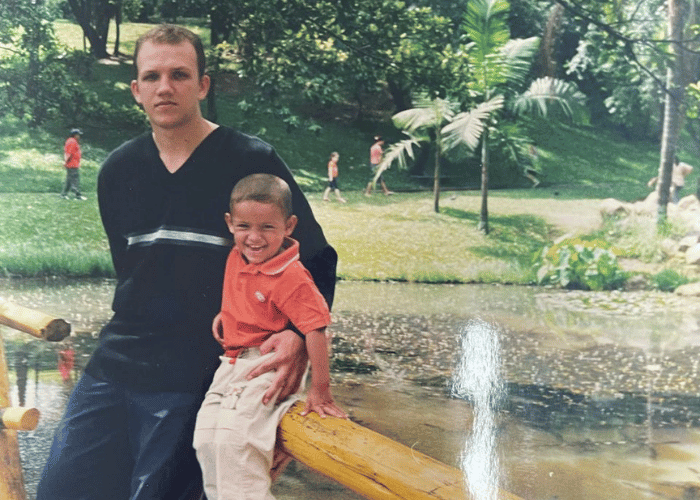 Foto antiga de pai e filho sorrindo ao lado de pequeno lago com fonte em parque arborizado, criança vestindo camisa laranja e adulto de camisa preta.