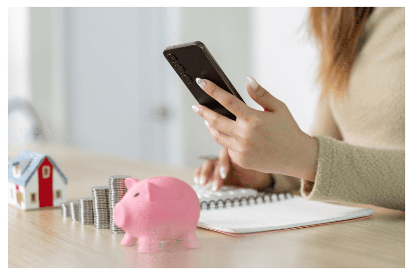 Pessoa sentada à mesa segurando um celular enquanto anota em um caderno. À frente, há um cofrinho rosa em forma de porco, pilhas de moedas e uma miniatura de casa, simbolizando planejamento financeiro e economia pessoal.