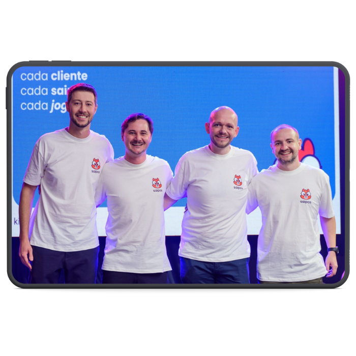 imagem mostra um tablet com os 4 fundadores, sendo o fundo composto por uma tela projetada em tom azul, parte do mascote da Saipos, o esquilo Allen, e os dizeres "impulsionar cada cliente"