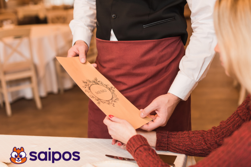 garçom com uniforme preto, branco e marrom entrega cardápio bege para cliente sentada à mesa. Ela é loira, com camiseta bordô