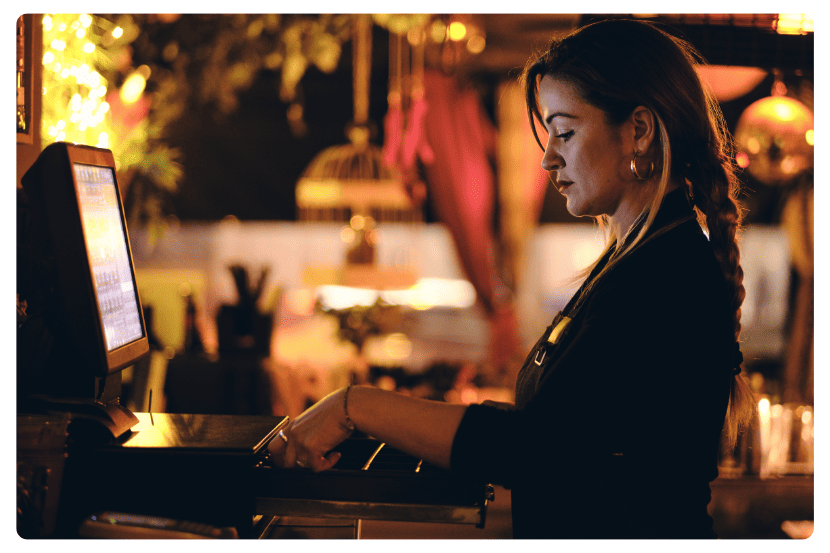 Mulher em um restaurante abrindo a gaveta do caixa para realizar uma sangria de caixa. Ela está concentrada, usando uniforme escuro, com um computador de frente e iluminação amarelada ao fundo, típica de ambiente de restaurante.