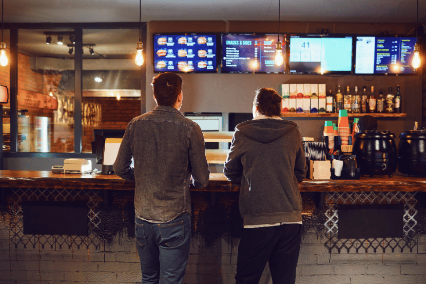 Dois clientes em frente ao balcão de atendimento de restaurante fast food, observando painéis digitais de menu iluminados acima, com área de bar e copos coloridos à direita