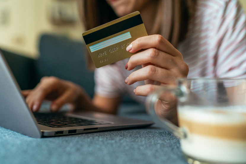 mulher segurando cartão de crédito em frente a um notebook. Ao seu lado uma xícara de capuccino.